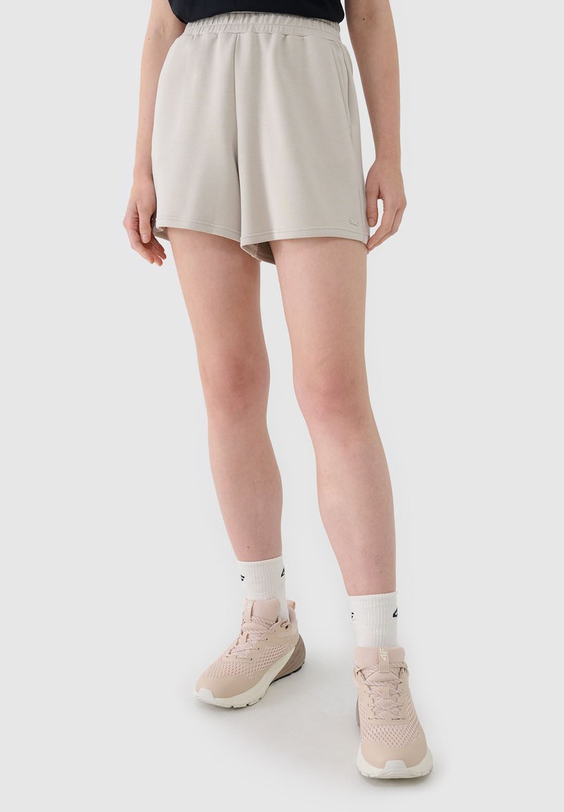 Femme portant un short beige à taille élastique, des chaussettes blanches montantes et des chaussures de sport rose clair, debout devant un fond clair uni.