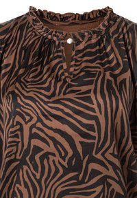 Bluse mit Zebra-Print in Braun und Schwarz, rüschigem Ausschnitt, Schlüsselöffnungsdesign und weicher Stofftextur. Präsentiert auffällige wellenartige Muster.