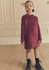 Robe tricotée magenta scintillante avec des manches longues et un col haut, associée à des bottes noires à lacets et des chaussettes noires de longueur cheville.