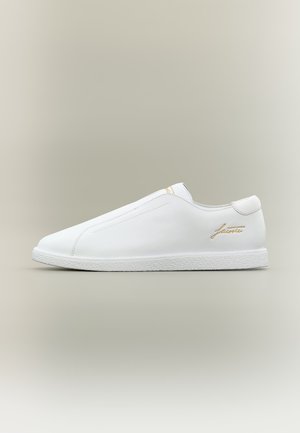 Witte instapsneaker met een textuur zool en een goudkleurig "Lacoste" logo aan de zijkant, getoond tegen een neutrale achtergrond.