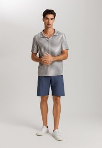 Polo a righe bianco e blu navy, abbinato a pantaloni corti blu. Modello che indossa sneakers bianche, con le braccia incrociate, in piedi davanti a uno sfondo neutro.