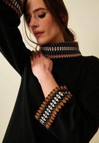 Nainen koskettaa mustan neuleen kaulusta, jossa on oransseja, valkoisia ja harmaita geometrisia kuvioita kauluksessa ja hihansuissa, katsoen alaspäin.