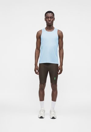 Mand iført en lyseblå ærmeløs atletisk top, mørkebrune tætsiddende shorts, hvide sokker og hvide sneakers, stående mod en ensfarvet baggrund.