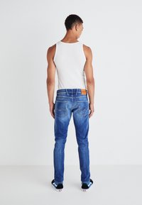 Jeans in denim blu con una vestibilità slim, finitura sbiadita e cuciture distintive sulle tasche posteriori; indossati con una canotta bianca semplice.