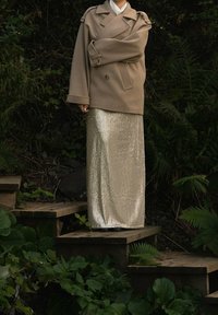 Beżowy, dwurzędowy wełniany płaszcz z oversize'owymi rękawami, zestawiony z długą, błyszczącą złotą spódnicą z cekinów, stojący na drewnianych schodach pośród zieleni.