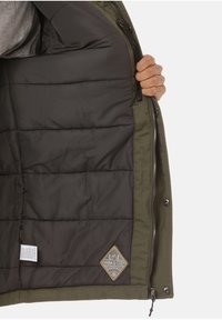 Mano sosteniendo una chaqueta verde abierta que muestra el interior acolchado negro con un parche que dice "Cálido y Acogedor" y una etiqueta de cuidado en el interior.