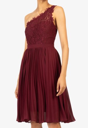 Mujer con un vestido burdeos hasta la rodilla con un hombro de encaje y una falda plisada, de pie frente a un fondo liso.