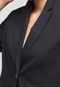 Dark navy blazer with a textured fabric, featuring a sharp lapel, single button closure, and a fitted silhouette, showcasing clean lines.