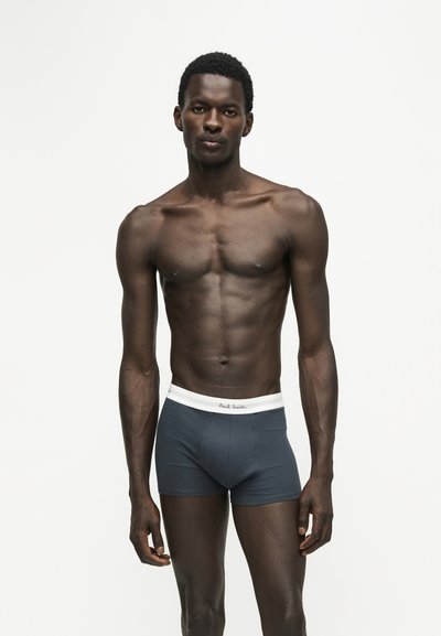 Homme mince à la peau foncée portant un boxer bleu foncé avec une ceinture blanche, debout devant un fond clair uni.