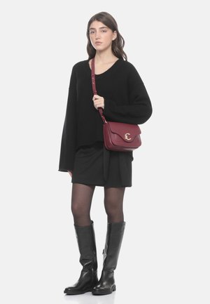 Donna che indossa un maglione nero oversize, gonna nera, collant neri, stivali alti neri e porta una borsa a tracolla di pelle bordeaux.