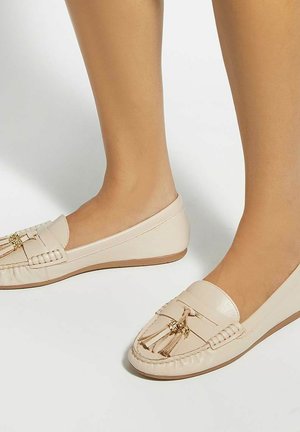 Voeten met beige leren loafers met kwastjes, staand op een lichte ondergrond.