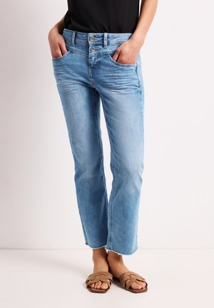 Kvinde iført lyseblå korte jeans med dobbelte knapper og brune flettede åbne sandaler, hænderne i lommerne.