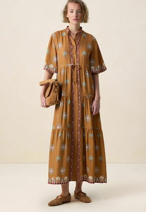 Femme portant une longue robe moutarde avec des boutons, des motifs géométriques et floraux, tenant un sac marron et chaussant des ballerines marron cloutées.