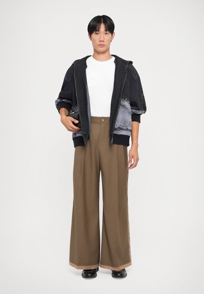 Black hoodie with gray accents, white t-shirt, and wide brown trousers with a side stripe. Model holding a small black wallet.