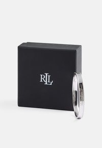 Pulsera de brazalete de plata con una superficie lisa, borde texturizado y detalles grabados. Presentada junto a una caja negra con un logo blanco.