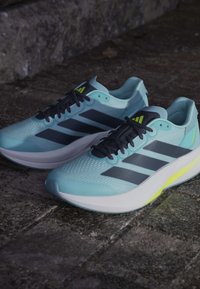 Baskets de course bleu clair avec des rayures marine et des lacets noirs. Elles possèdent une tige texturée, une semelle rembourrée et un accent jaune vif au niveau du talon.