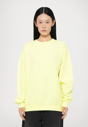 Bright yellow oversized sweatshirt with long sleeves, ribbed cuffs, and a crew neck, worn over dark jeans. Smooth, soft fabric texture.