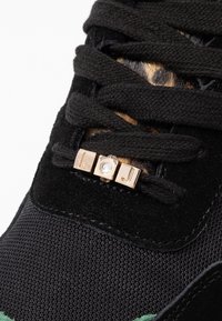 Svarta sneakers med strukturerad svart textil och mocka, prydda med guldfärgad hårdvara och en klar sten på snörningen.