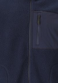Marinblå fleecejacka med en texturerad yta, en ficka med dragkedja på bröstet och sidofickor för händerna, samt en slät tygpanel på övre framsidan.