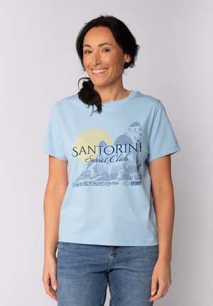 Lachende vrouw met donker haar in een paardenstaart, gekleed in een lichtblauw T-shirt met de tekst "Santorini Sunset Club" en een blauwe spijkerbroek, staand tegen een witte achtergrond.