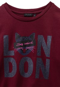Bordeaux katoenen T-shirt met glinsterende, donkere blauwe letters die "LONDON" spellen en een afbeelding van een kat met de tekst "Stay weird" in het roze.
