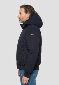 Marineblaue Steppjacke mit großem Kapuzen, Reißverschluss, Seitentaschen und gerippten Bündchen. Verfügt über ein Logo-Patch am Oberarm.