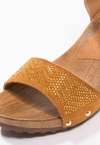 Sandale en daim marron avec une large sangle embellie de clous dorés, posée sur une semelle en bois texturé de couleur marron clair.