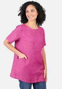 Pink linen tunic top with short sleeves, round neckline, side pockets, and a relaxed fit, paired with blue jeans. Smooth texture and lightweight.