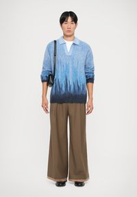 Maglione a maglia con gradiente blu e colletto polo, indossato sopra una camicia bianca. Pantaloni larghi marroni a gamba larga e scarpe nere. Borsa nera semplice.
