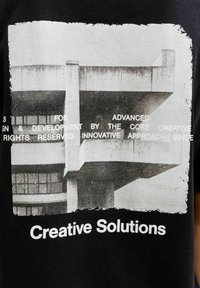 Czarna koszulka z monochromatyczną grafiką betonowej konstrukcji oraz napisem "Creative Solutions" na dole.