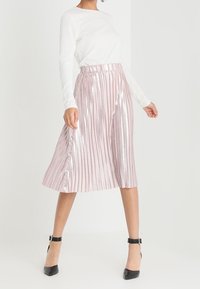 Plissierter Midirock in metallischem Rosé mit hoher Taille und elastischem Bund. Kombiniert mit einem weißen Langarmshirt und schwarzen Absätzen.