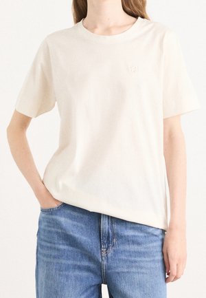 Roomkleurig T-shirt met korte mouwen van zacht katoen, met een ronde halslijn en een subtiel reliëflogo op de linkerborst. Draagt bij blauwe spijkerbroek.