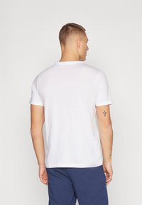 Uomo visto di spalle che indossa una semplice maglietta bianca a maniche corte e pantaloni blu scuro, con un tatuaggio a forma di croce sul braccio sinistro.