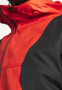 Červená a černá bunda s kapucí na zip, s kontrastními panely, hladkou texturou a viditelnými švy na tkanině.