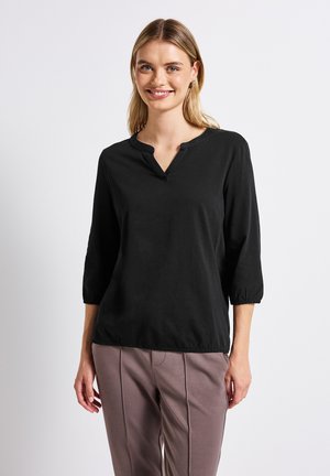 Haut noir léger avec un col en V, des manches trois-quarts froncées aux poignets et une coupe décontractée. Porté avec un pantalon marron clair.