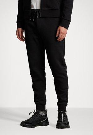 Personne portant un pantalon de jogging noir avec une taille à cordon et des chaussures de sport noires avec des chaussettes à motifs, debout devant un fond blanc.