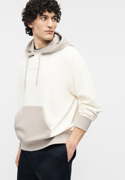 Νεαρός άνδρας με σγουρά μαλλιά που φοράει μια μπλούζα με κουκούλα σε κρεμ και μπεζ αποχρώσεις με μπροστινή τσέπη, κοιτάζει προς τα πλάγια, με τα χέρια εν μέρει στις τσέπες ενός μαύρου παντελονιού.