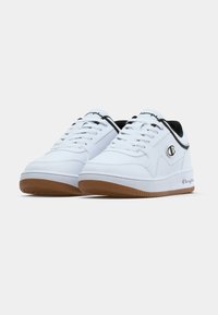 Białe buty sportowe z gładką skórzaną powierzchnią, czarnymi akcentami, jasnoszarym wnętrzem i brązową gumową podeszwą. Na boku znajduje się emblem logo.