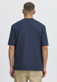 Tricou din bumbac bleu marin, cu o croială lejeră, guler rotund, mâneci scurte și detalii de cusătură vizibile, purtat cu pantaloni khaki.