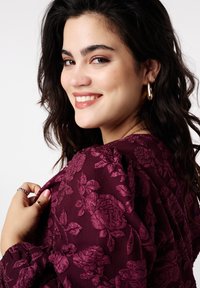 Glimlachende vrouw met donker krullend haar, gouden creolen en een donkerpaarse top met bloemenpatroon, die de stof bij haar schouder vasthoudt.