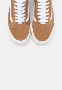 Baskets en daim marron avec une semelle en caoutchouc blanc, dotées de lacets blancs et de rayures latérales. Texture lisse avec un bout arrondi et des détails cousus.