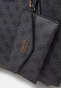 Sort taske med et tekstureret stof, der har et subtilt mønster. Guldfarvet "GUESS" logo er synligt. En matchende pung er fastgjort til tasken.
