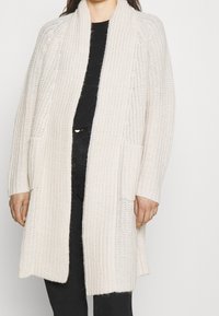 Kramfärgad lång cardigan i grovstickad kvalitet, med sjalkrage, öppen front och två framfickor.