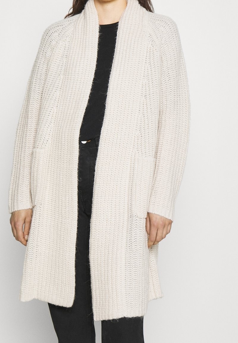 Kramfärgad lång cardigan i grovstickad kvalitet, med sjalkrage, öppen front och två framfickor.