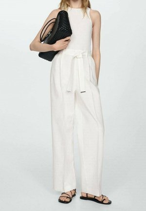 Kobieta w białym, bezrękawnym kombinezonie z wiązanym paskiem w talii, trzyma czarną plecioną torebkę i nosi czarne sandały na ramiączkach.