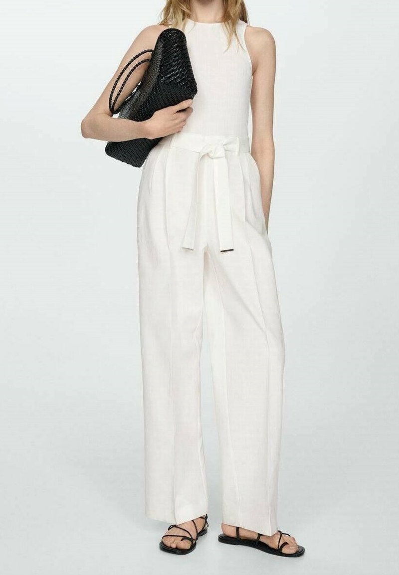 Femme portant une combinaison blanche sans manches avec ceinture nouée à la taille, tenant un sac à main tressé noir et portant des sandales noires à fines sangles.