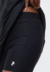 Άτομο τραβάει προς τα πάνω μαύρα αθλητικά σορτς με ένα μικρό λευκό λογότυπο στο αριστερό μηρό σε μονόχρωμο φόντο.