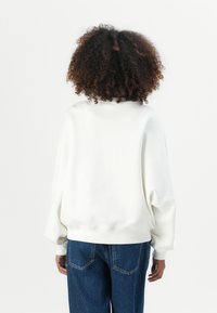 Felpa bianca oversized con scollatura rotonda, spalle cadenti e polsini a coste, abbinata a jeans in denim blu. Texture liscia.