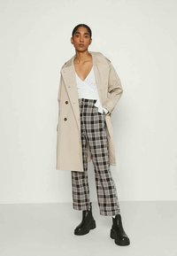 Trench coat bege com grandes botões, combinado com calças de cintura alta em padrão xadrez preto e branco e botas de tornozelo pretas chunky.