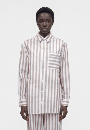 Osoba stojaca v béžovej a bielej vertikálne pruhovanej gombíkovej košeli so zodpovedajúcimi nohavicami, pruhy na vreckách na hrudi sú horizontálne.